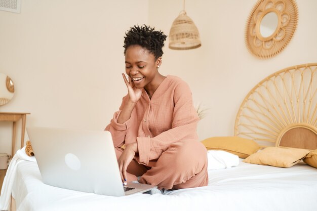 Heureuse jeune fille assise sur le lit dans la chambre et utilisant un pyjama en mousseline pour ordinateur portable discutant avec des amis