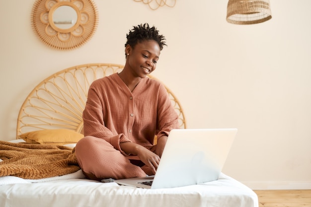 Heureuse jeune fille assise sur le lit dans la chambre et utilisant un pyjama en mousseline pour ordinateur portable discutant avec des amis