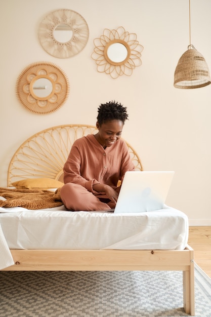 Heureuse jeune fille assise sur le lit dans la chambre et utilisant un pyjama en mousseline pour ordinateur portable discutant avec des amis