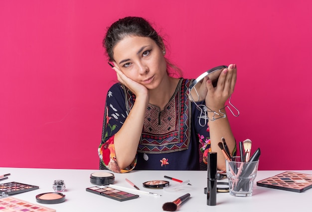 Photo heureuse jeune fille brune assise à table avec des outils de maquillage tenant un pinceau de maquillage et un miroir