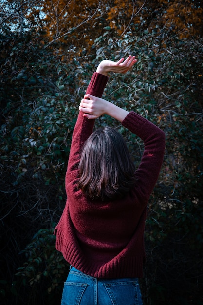 Heureuse jeune fille brune dans un pull marron debout avec levant les mains dans un paysage forestier