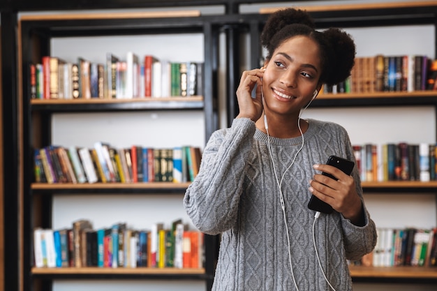 Heureuse jeune fille étudiante africaine étudiant à la bibliothèque, écoutant de la musique avec des écouteurs