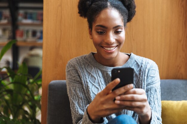 Heureuse jeune fille étudiante africaine qui étudie à la bibliothèque, à l'aide de téléphone mobile