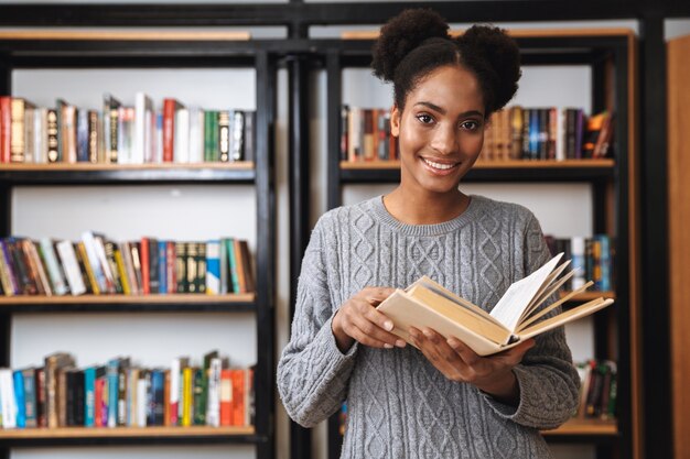 Heureuse jeune fille étudiante africaine qui étudie à la bibliothèque, lisant un livre