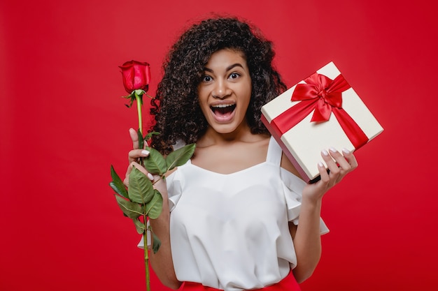 Heureuse jeune fille noire souriante avec rose et coffret cadeau isolé sur rouge