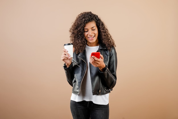 Heureuse jeune fille noire souriante avec téléphone et café pour aller tasse isolé sur marron