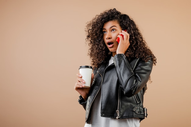 Heureuse jeune fille noire souriante avec téléphone et café pour aller tasse isolé sur marron