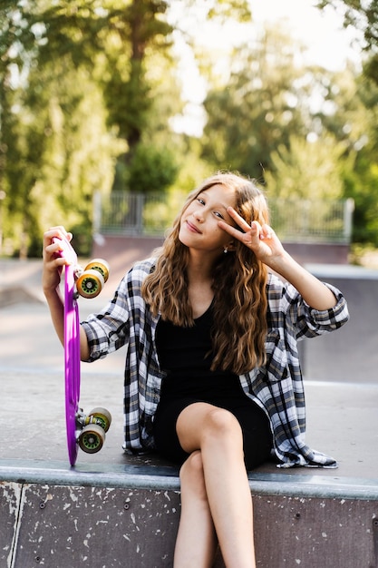 Heureuse jeune fille avec penny board assis sur une aire de skate et sourire Mode de vie sportif extrême Enfant souriant avec penny board posant sur une rampe de sport