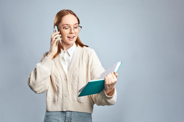 Heureuse jeune fille portant un pull décontracté parlant sur un téléphone portable tout en lisant un cahier