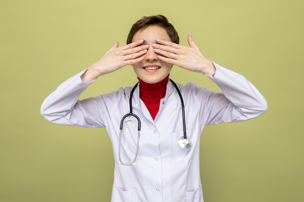 Heureuse jeune fille souriante médecin en blouse blanche avec stéthoscope autour du cou yeux coniques avec paumes debout sur vert