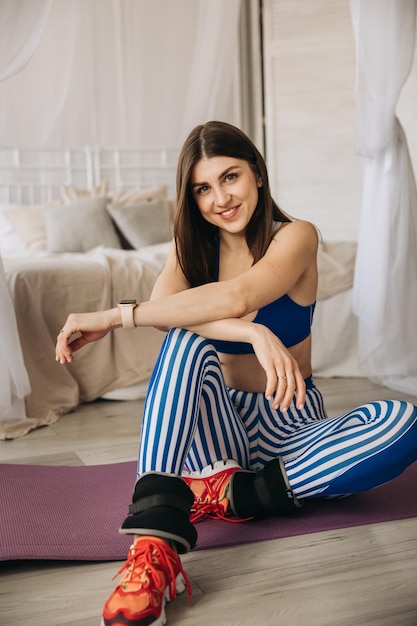 Heureuse jeune fille sportive et souriante est assise sur un tapis de yoga à la maison, faisant du sport à la maison en s'étirant haut ...