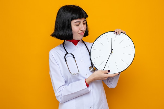 Heureuse jeune jolie femme caucasienne en uniforme de médecin avec stéthoscope tenant et regardant l'horloge