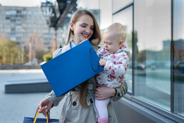 Heureuse jeune maman avec petite fille sur les bras et sacs à provisions à la main. Jour de shopping.
