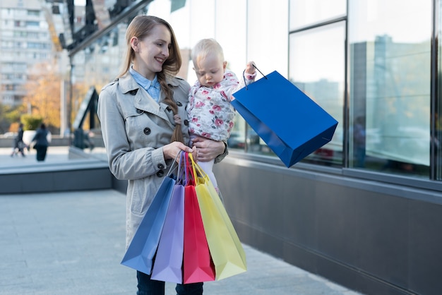 Heureuse jeune maman avec petite fille sur les bras et sacs à provisions à la main.