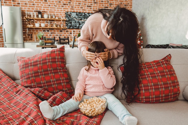 Heureuse jeune maman et petite fille rient en étant assise sur le canapé et mangeant du pop-corn. famille regardant la télévision et mangeant du pop-corn.