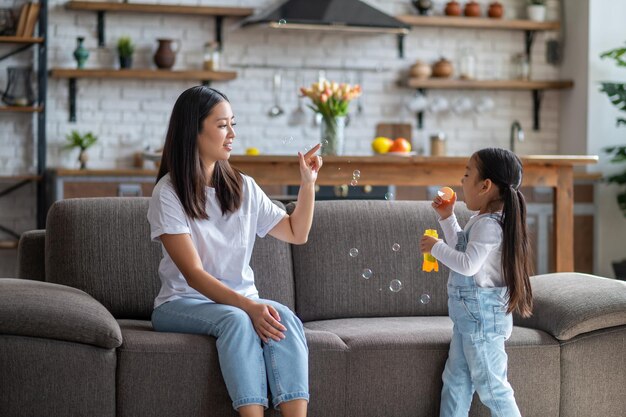 Heureuse jeune mère assise sur le canapé tout en faisant éclater les bulles que sa fille souffle avec la baguette