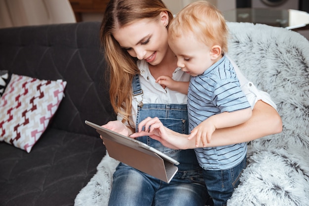 Heureuse jeune mère avec son petit fils assis et utilisant une tablette sur un canapé