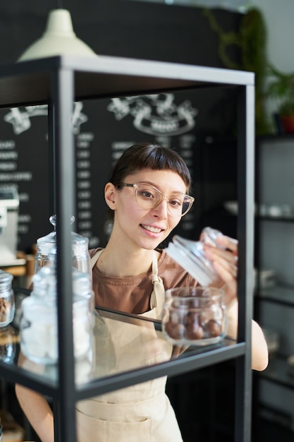 Heureuse jeune serveuse brune à lunettes et uniforme regardant dans un pot avec du thé