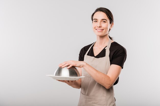 Heureuse jeune serveuse brune en tablier en gardant la main sur le couvercle de la cloche en se tenant debout devant la caméra dans l'isolement