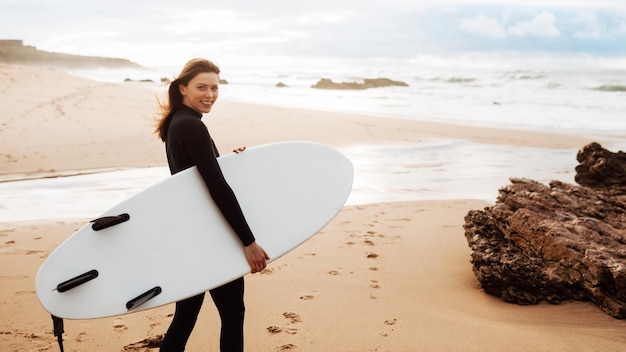 Heureuse jeune surfeuse marchant sur la plage avec sa planche de surf dans le panorama du matin avec