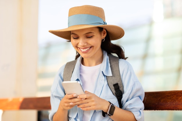 Heureuse jeune touriste européenne en chapeau avec sac à dos tapant sur un smartphone utilisant une carte en ligne pour discuter