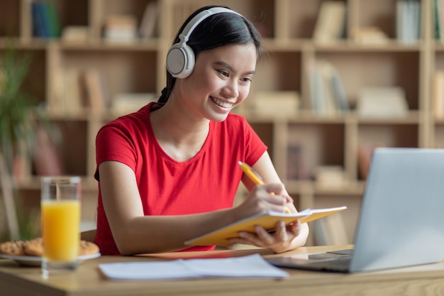 Heureuse jolie adolescente japonaise dans les écouteurs prend des notes pour écouter l'étude de la conférence sur un ordinateur portable à l'intérieur de la chambre