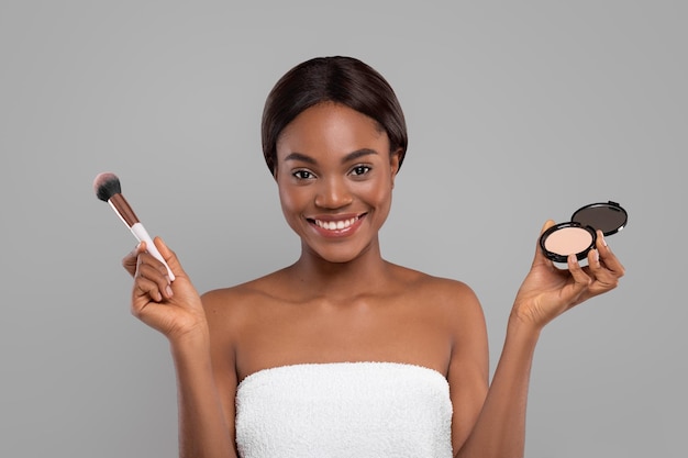 Heureuse jolie dame noire avec une peau parfaite faire un maquillage de base avec des cosmétiques tenir une poudre compacte et