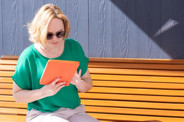 Heureuse jolie étudiante en lunettes de soleil travaillant à distance dans le parc à l'aide d'une tablette numérique