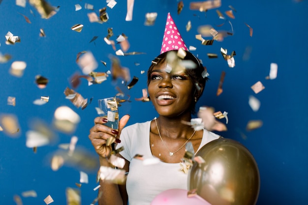 Heureuse jolie femme africaine en t-shirt blanc danse heureuse et jetant des confettis, fête son anniversaire. Photo intérieure de jolie dame noire tenant du champagne et des ballons avec une expression de visage heureux