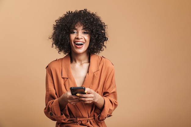 Heureuse jolie femme africaine tenant un smartphone et regardant directement sur fond marron