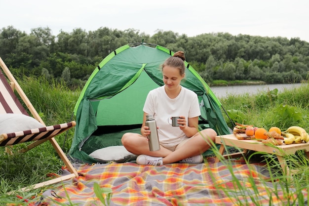 Heureuse jolie femme assise sur le sol près de la tente et de la rive ayant des loisirs et un voyage en plein air tenant un thermos et une tasse en dégustant une boisson chaude en plein air