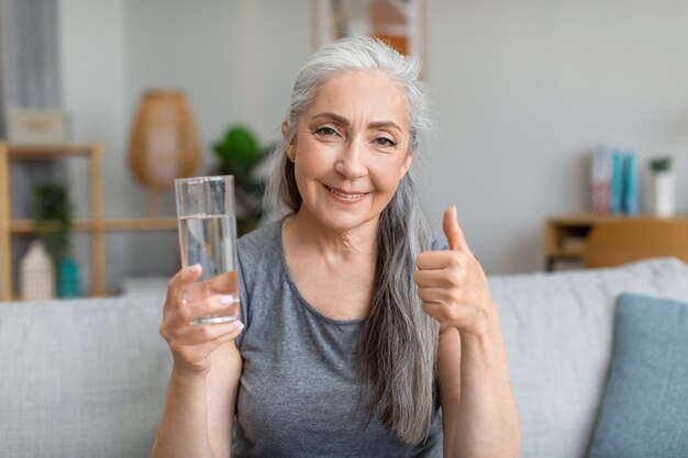 Heureuse jolie femme aux cheveux gris senior caucasienne tient un verre d'eau et montre le pouce vers le haut