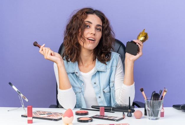 Heureuse jolie femme caucasienne assise à table avec des outils de maquillage tenant une tasse gagnante et un pinceau de maquillage isolé sur un mur violet avec espace de copie