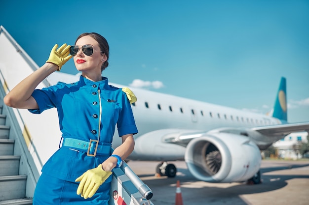 Heureuse jolie femme en uniforme élégant et lunettes de soleil se tient sur l'escalier de l'avion sur la piste par une journée ensoleillée