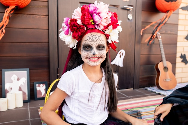 Heureuse jolie fille au visage peint assis devant la caméra contre le porche et la porte de la maison décorée de symboles traditionnels d'halloween