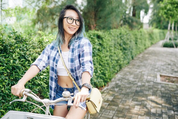 Heureuse jolie jeune femme chinoise à lunettes à vélo dans le parc de la ville