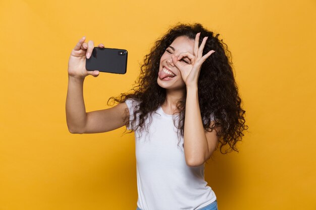 Heureuse jolie jeune femme posant isolée sur jaune prendre un selfie par téléphone mobile.