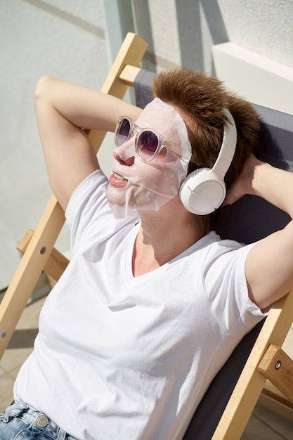 Heureuse jolie jeune fille avec un masque en tissu sur le visage assis sur le balcon et écouter de la musique sur les écouteurs.