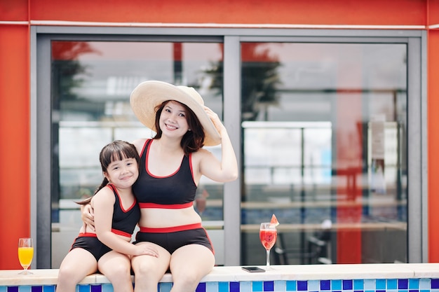 Heureuse jolie petite fille serrant sa mère dans un grand chapeau de paille quand ils se reposent à la piscine de l'hôtel spa