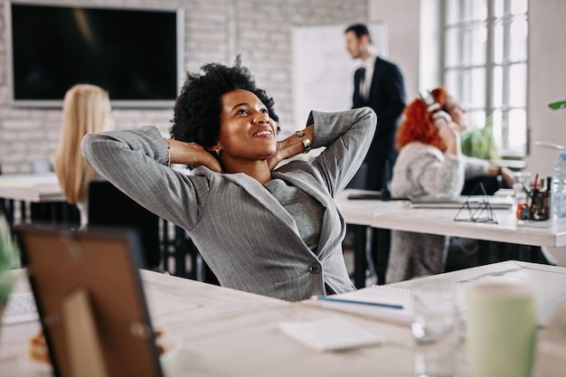 Heureuse journée de femme d'affaires noire rêvant assise à son bureau avec les mains derrière la tête et se relaxant Il y a des gens en arrière-plan