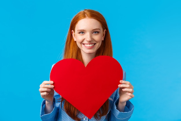 Heureuse, joyeuse et charmante, tendre femme de race blanche tenant une grande carte de Saint Valentin coeur rouge et souriant, donnant son amour et ses soins, félicite son petit ami avec leur rendez-vous romantique, mur bleu