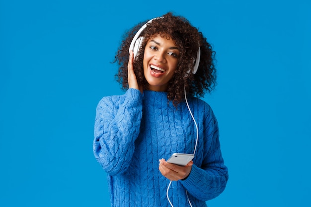 Heureuse et joyeuse femme afro-américaine jouant à l'application de jeu de karaoké dans un smartphone, portant des écouteurs chantant et appréciant une musique impressionnante, une qualité sonore, un fond bleu debout