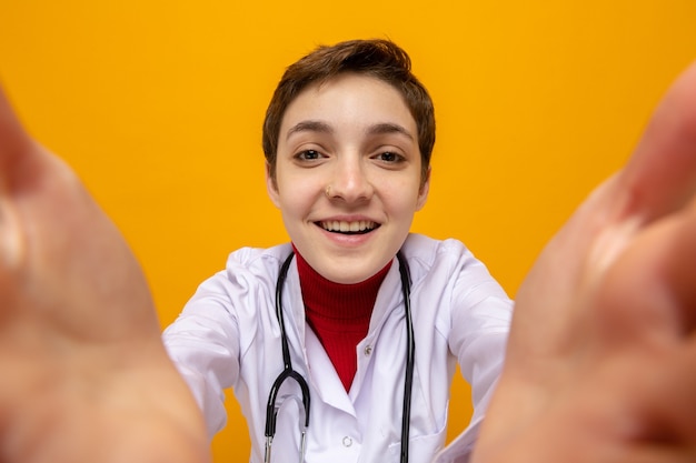 Heureuse Et Joyeuse Jeune Fille Médecin En Blouse Blanche Avec Stéthoscope Autour Du Cou Prenant Selfie Regardant Devant Souriant Debout Sur Un Mur Orange