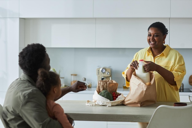 Heureuse maman afro-américaine sortant des sacs d'épicerie et parlant à sa famille dans la cuisine