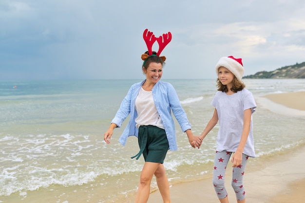 Heureuse maman et fille enfant en bonnet de Noel marchant main dans la main le long de la plage
