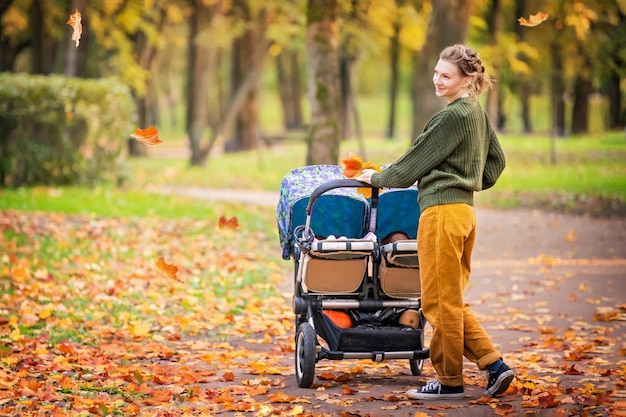 Photo heureuse maman marchant dans le parc d'automne avec une poussette pour jumeaux