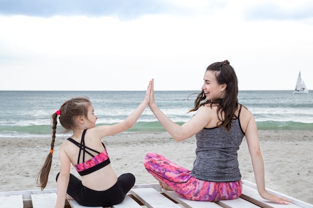 Heureuse maman et sa fille en vêtements de sport pratiquent le yoga au bord de la mer. Des valeurs familiales et un mode de vie sain.