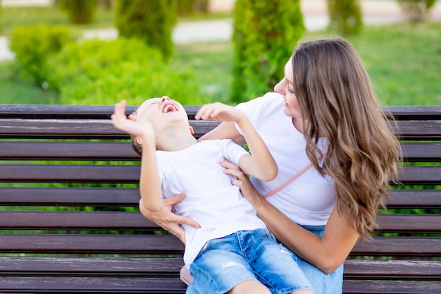Heureuse maman avec son bébé dans le parc sur un banc en été s'amuser à se serrer dans ses bras, rire et jouer