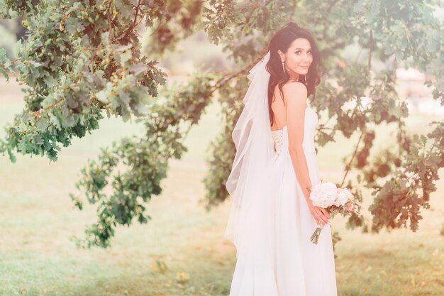 Heureuse mariée bronzée dans une belle robe blanche posant avec bouquet de fleurs en journée ensoleillée.