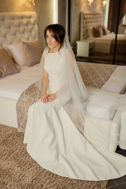 Heureuse mariée dans une robe de mariée est assise sur un lit dans un hôtel. Matin de mariage de la mariée.